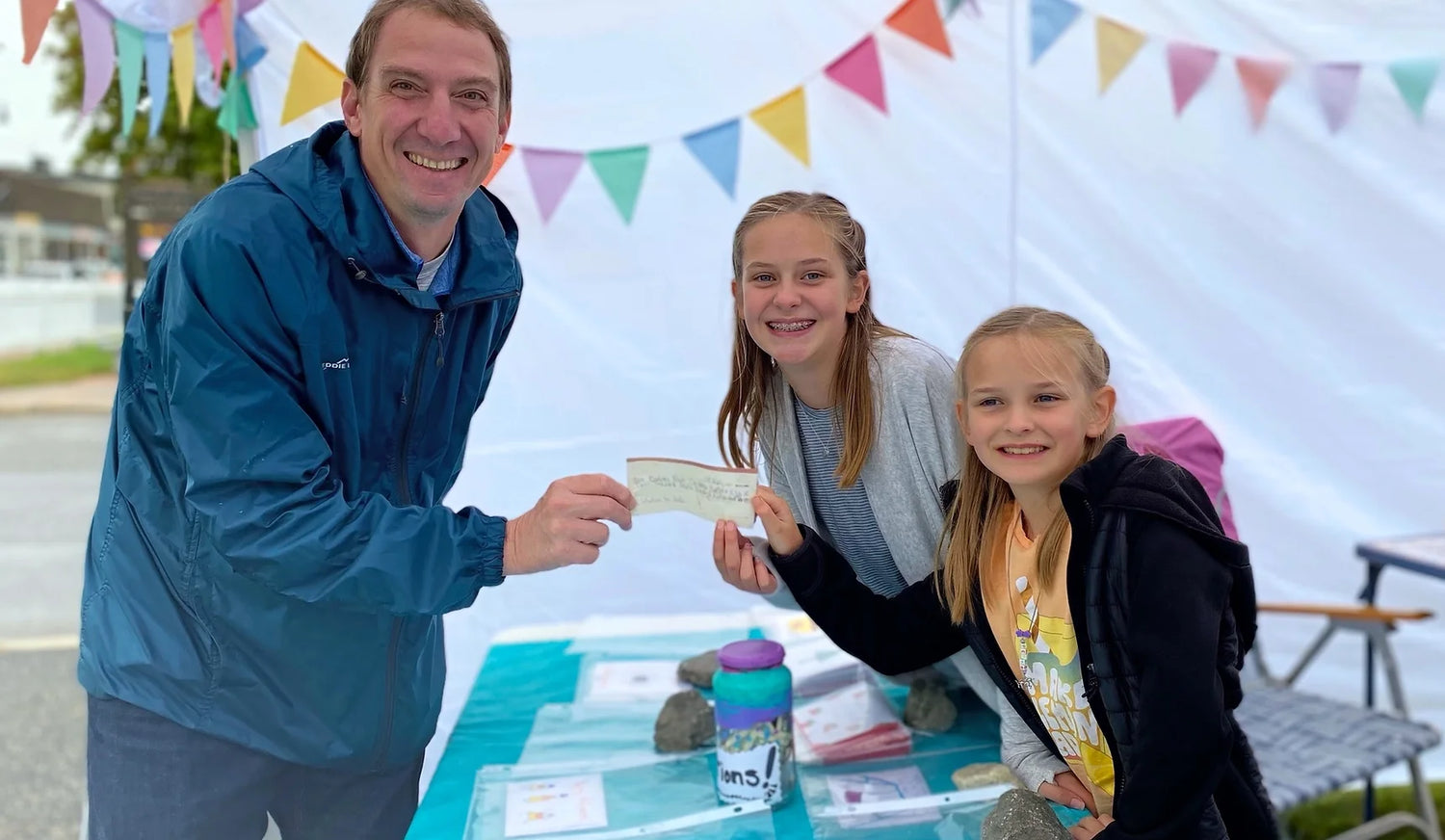 The sisters handing a donation check to an adult, in a marketplace setting.